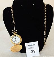 Gold tone pocket watch open to show white dial with black numbers and ornate hands, hanging on black display stand with attached chain.