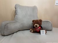 Adult size gray reading pillow with arm supports and backrest, accompanied by a brown teddy bear holding a heart.