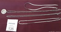 Four silver chains displayed on fabric background: a 16-inch serpentine chain, a 25-inch paperclip chain with Mexican pendant, a 24-inch multi-link chain, and an 18-inch braided serpentine chain.