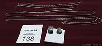 Four silver chains laid out along with one pair of silver 'X' earrings on a dark surface, with lot tag number 138.