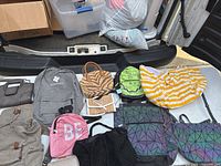 Photo showing a variety of 14-15 purses, bags and backpacks arranged on a vehicle surface. Items include a grey Osprey backpack, two holographic geometric tote bags, a large yellow and white striped soft bag, a black fabric bag, a pink small backpack with 'BE' letters, a zebra-pattern handbag, a brown/tan backpack, and several small purses of assorted colors.