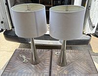 Two 2 ft tall table lamps side by side on wooden surface, showing polished metal tapered bases and white fabric drum shades.