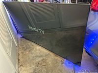 Front view of the 70-inch Elements Roku TV placed on the floor against a wall showing screen intact but reflective.