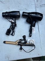 Photo showing two black hair dryers and one gold curling wand placed on a light-colored surface. All items have attached power cords and visible branding 'HOT BEAUTY'.