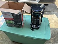 Overall view of the Black & Decker coffee maker placed on a green surface with its original box nearby showing name and features.