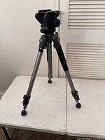 Front angle view of silver aluminum camera tripod with black pan-tilt head and three legs with locking clamps. Visible missing screw on bottom of one leg.