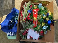 Box with assorted Christmas decorations including lighted garland, ornamental baubles, and stockings alongside a blue bag potentially containing lights.