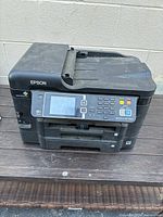Front and top angle view of the black Epson Workforce WF-3640 all-in-one printer on a wooden surface with visible dust and dirt.