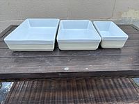 Three white rectangular glass cooking trays side by side on a dark wood surface showing their heights and stackable form.