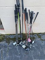 Photo showing 25 mixed golf clubs standing upright against a white wall on rough pavement including drivers, irons, and putters with mixed shaft and grip colors.