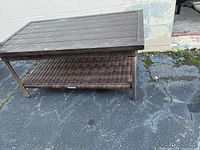 Full view of the brown outdoor side table showing wood plank top, metal legs, and woven rattan lower shelf on asphalt surface.