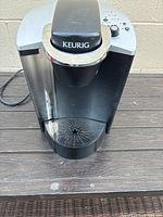 Front view of the black and silver Keurig coffee maker showing the pod slot and drip tray with star-shaped holes.