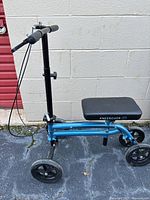 KneeRover blue knee walker with padded knee platform, four wheels, and handlebar brakes against a concrete block wall.