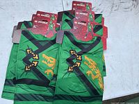 Six green fabric costume tops stacked showing identical Ninjago ninja design with yellow dragon and black straps, with red Lego Ninjago label tags for ages 6+.