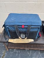 Blue Porta Brace camera bag with black front pocket and beige shoulder strap showing wear, placed on a brown bench outdoors.