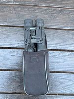 Front view of Tasco MX33 binoculars in black, resting on a wooden surface with carrying case underneath.