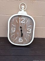 Front view of white oval decorative wall clock with wood effect face showing numerals 12, 3, 6, 9 and black hands