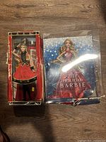 Photo showing two Barbie dolls in original packaging on wooden floor. Boxes have visible wear and damage, but dolls appear intact and well-preserved inside.