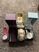 Three wristwatches displayed on a carpeted floor with their boxes showing. Pink, white rectangular, and silver Bangle watches.