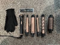 Photo of the entire curling set showing the main curling iron handle, six interchangeable rose gold curling wands, a black heat-resistant glove and a small plastic bag with hair clips.