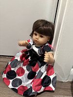 Full view of the reborn doll sitting, showing fabric polka dot dress with red, black, and white dots and black ribbon
