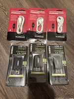 Three NUMOJO white micro USB cables in packaging and three Uniden USB AC chargers in packaging arranged on floor.