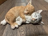 Front angled view of the resin figurine showing orange and white tabby cat cuddling a grey and white cat, both sleeping.