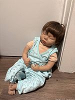 Reborn doll seated against a white wall, showing full body in light blue outfit with floral pattern and peach inner shirt. Doll has realistic vinyl limbs and closed eyes, brown hair neatly styled.