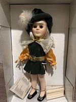 Full frontal view of Prince Charming doll dressed in green velvet outfit with orange sleeves, white lace collar and feathered hat, with accessories in box