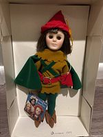 Full view of Effanbee Robin Hood doll in original box wearing green and brown costume with red belt and shoes, with red hat accessory positioned on the head. Original tag visible.