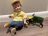 Porcelain doll sitting on floor next to John Deere tractor model showing both items.