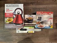Black and Decker electric kettle in box, Betty Crocker sandwich grill in box, MyMini noodle cooker and skillet box, and Mario Lopez grater and zester laid out on table. Shows overall lot contents.