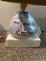 Lampshade showing lighthouse and coastal scene in blue tones, wrapped in plastic, placed on white foam base.