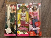 Three Barbie dolls in clear packaging, visible wear on boxes, including Holiday Joy, Easter Flowers, and a third Barbie with heart accessories.