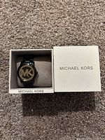 Black metal Michael Kors fashion watch in original white Michael Kors branded box on carpeted surface.