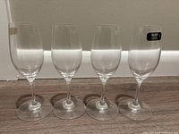 Photo showing four clear crystal stemmed spirit glasses with slender tapered bowls, arranged in a row against a beige wall and wooden floor baseboard.