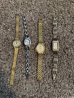 Photo showing four vintage analog wristwatches laid flat on a carpeted surface, displaying their faces and metal link bands in varying gold and silver tones.