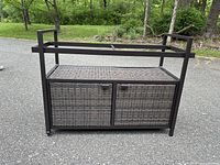 Front view of brown and black wicker and metal bar cart with two cabinet doors and caster wheels on asphalt driveway.