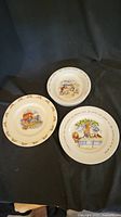 Three child's collector plates arranged on dark background: one Bunnykins bone china plate and two Alice in Wonderland ironstone plates