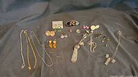 Overview of lot showing multiple gold and silver chains, earrings of various styles, a black enamel brooch, and a silver-tone lipstick case with tassel.