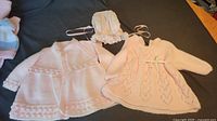 Two pink knitted sweaters and a white bonnet laid flat.