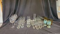 A set of 32 assorted drinking glasses of various styles and colors arranged in rows. Includes clear square water glasses, juice glasses, clear beer steins with handles, tall highball glasses, turquoise goblets, and amber-colored ribbed glasses.