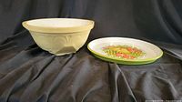 Ceramic mixing bowl and plastic fruit motif serving tray side by side on black cloth background.