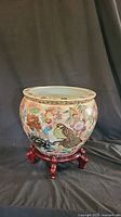Full view of the large round Chinese porcelain pot with floral and bird designs sitting on the wooden stand with five legs.