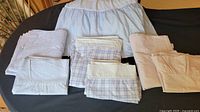 Photo showing folded bedding items including blue and white patterned sheets and dust ruffle, blue plaid sheets, and pink gingham sheets.