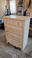 Front and side angle showing the 4-drawer chest in antiqued white with brass handles and floral detail on top drawer.