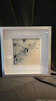 Front view of the framed modern art photographic print depicting abstract blue droplets and splatters with a white wooden frame.