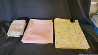 Three folded items arranged side by side on a black surface: taupe pillowcases, soft rose tablecloth, and yellow placemats with bird motifs.