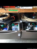 Shelf with four coiled extension cables atop drawer organizers containing junction boxes and bins of staples