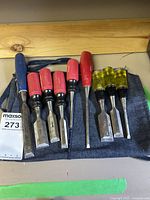 Nine chisels laid out in a denim roll with blue, red, and yellow handles set on a wooden surface
